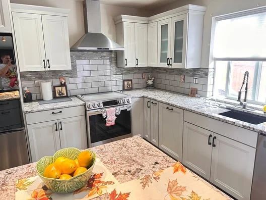 Bright kitchen with white cabinets, gray backsplash, stainless stove, and fruit bowl on a floral tablecloth.