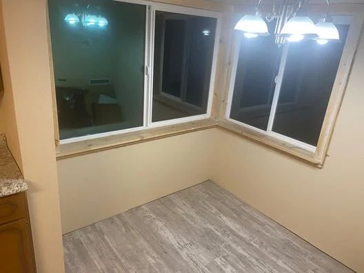 Empty corner nook with beige walls, gray wood-look floor, and large windows under hanging lights.