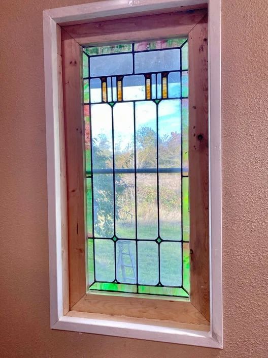 Small stained-glass window in a wooden frame on a pink wall with trees visible outside