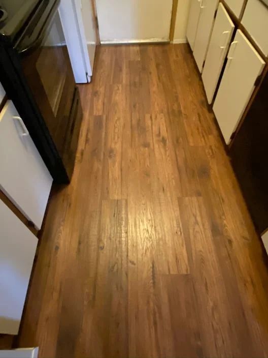 Narrow kitchen aisle with wood flooring, white cabinets on one side, and dark appliances on the other.