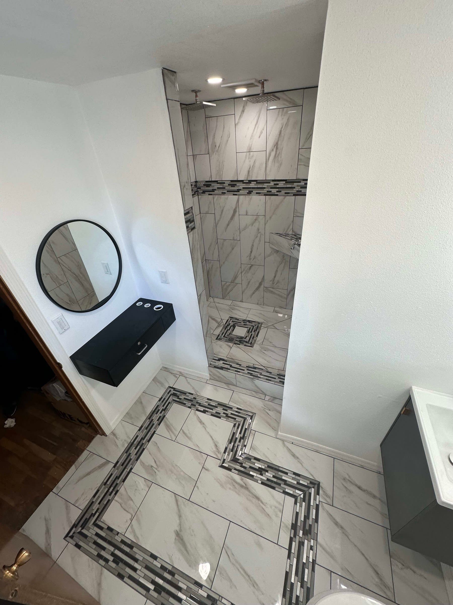 Small bathroom with marble-pattern tile shower, floor, sink, round mirror, and dark countertop.