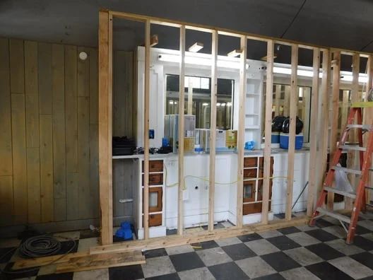 Partially framed kitchen renovation with exposed studs, white cabinets, and a red ladder on a checkered floor