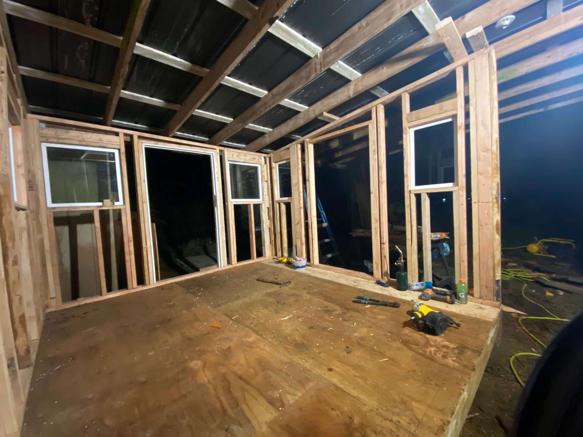 Unfinished wooden room interior with exposed framing, windows, and a plywood floor under construction at night