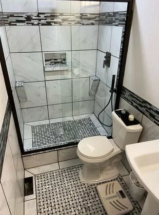 Small bathroom with tiled walk-in shower, toilet, and sink with a toilet-seat elevator assist device