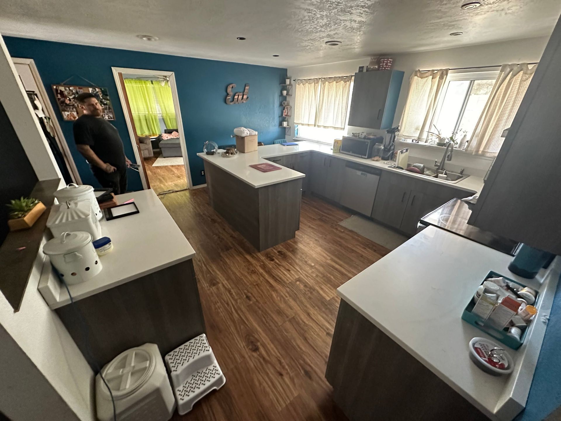 Bright kitchen with blue walls, white countertops, wood floor, and a person standing near the left doorway.