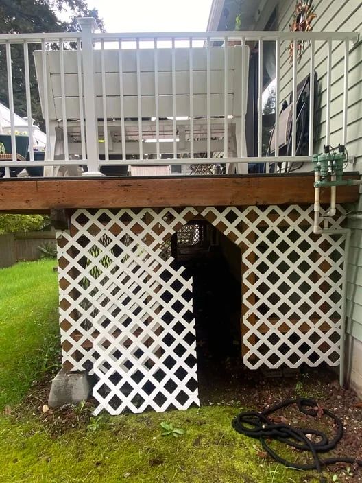 White lattice under raised deck with dark opening, gray railing above, and green lawn with black hose.
