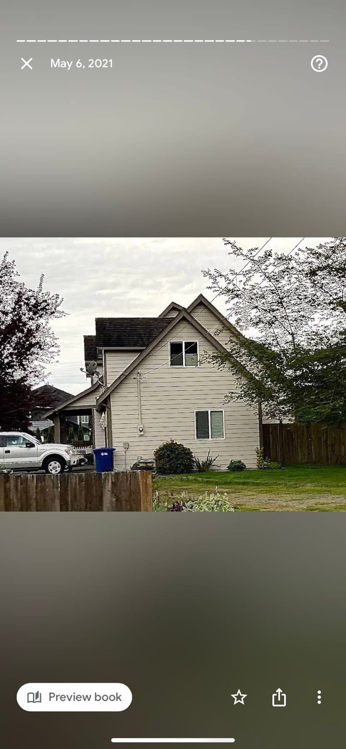 Small beige house with a steep roof, parked car, and trees under an overcast sky