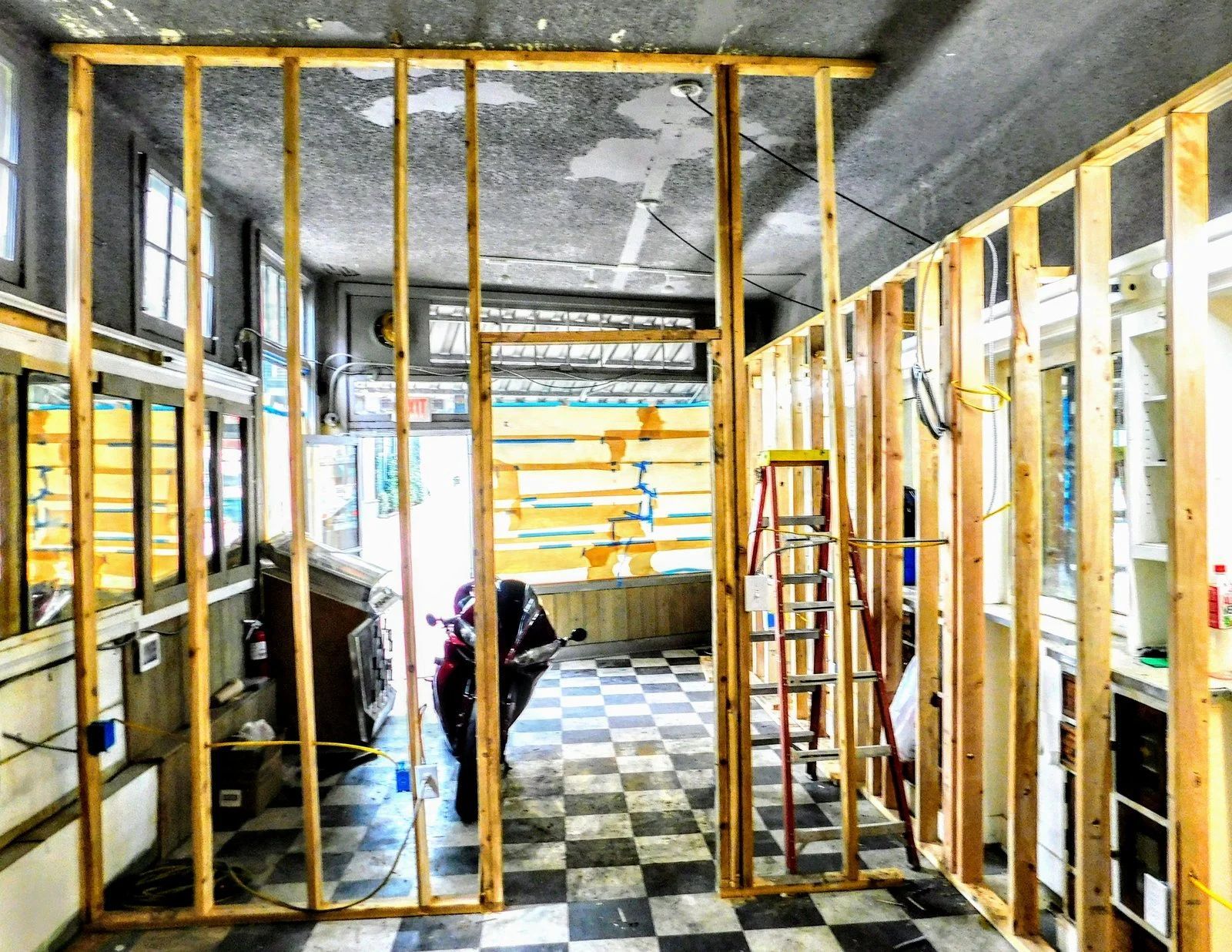 Interior of a house under renovation with exposed studs, checkered floor, and workers near the back wall