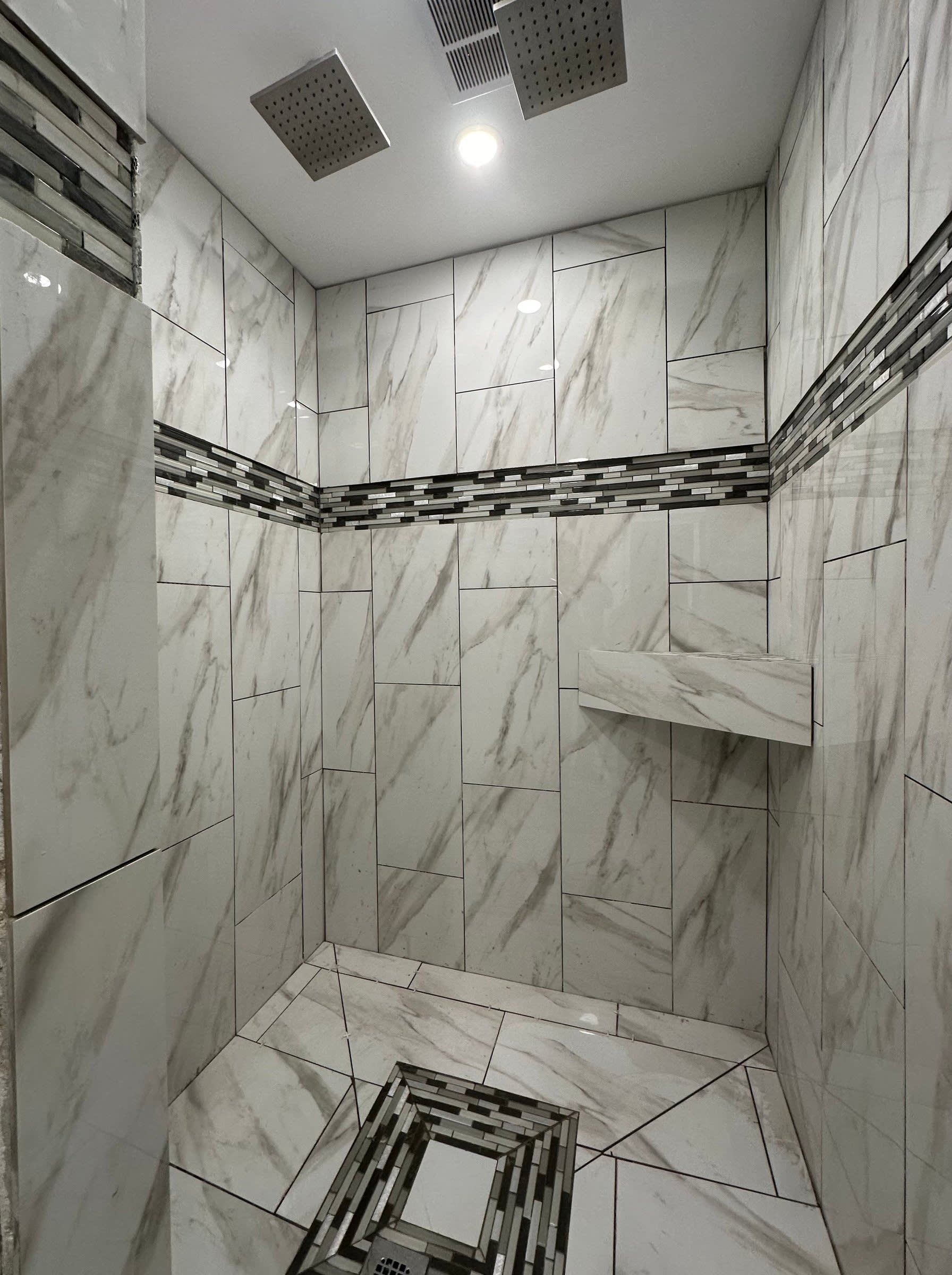 Modern tiled shower stall with white marble-look walls, dark accent band, ceiling light, and floor drain