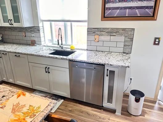 Kitchen sink and dishwasher under a window, with white cabinets, granite counters, and a trash can nearby.