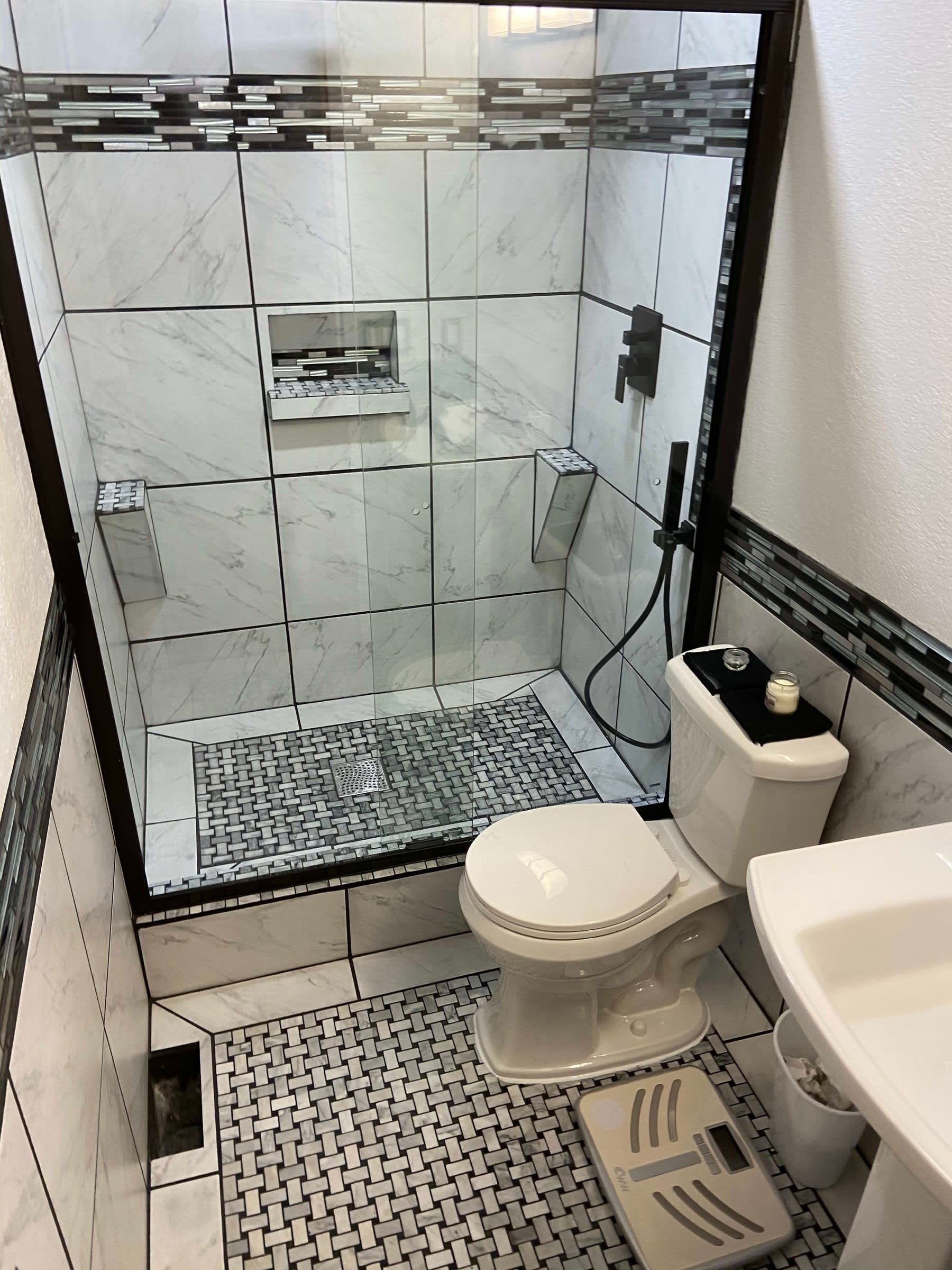 Small bathroom with glass shower, toilet, sink, and patterned floor mat.