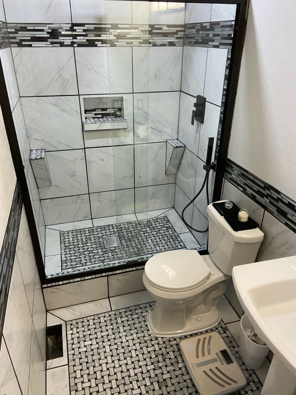 Small tiled bathroom with walk-in shower, toilet, sink, and patterned floor mats
