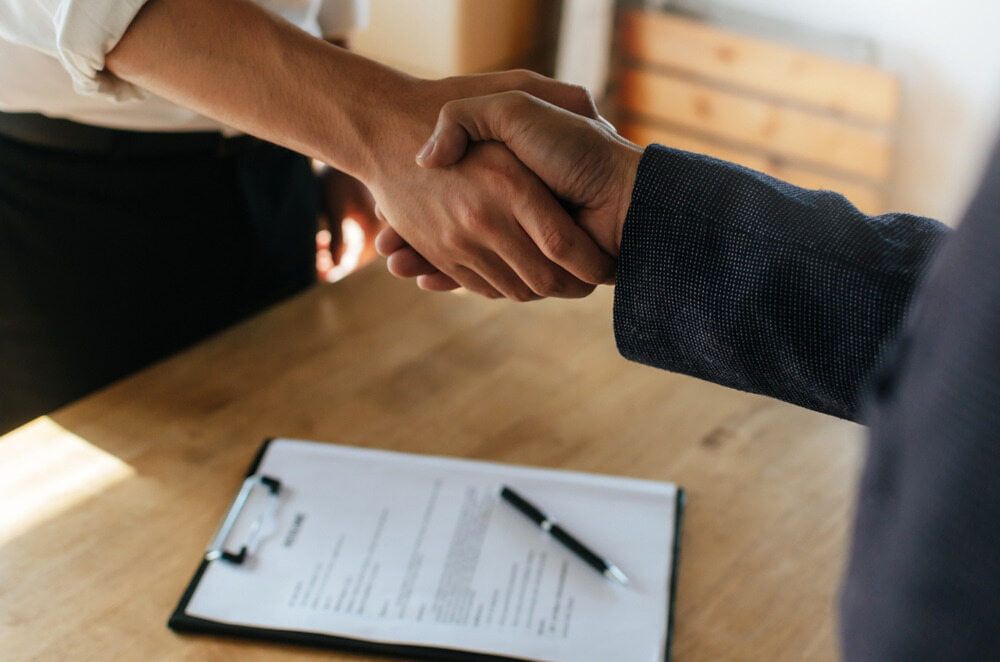 Two Men Are Shaking Hands Over a Clipboard with A Pen on It — Linda Emery & Associates In Gosford, NSW