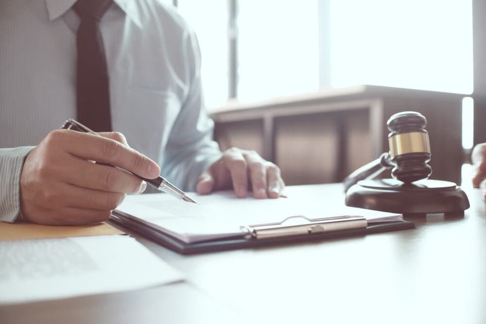 A Man Is Sitting at A Desk Writing on A Clipboard Next to A Judge 's Gavel — Linda Emery & Associates In Gosford, NSW