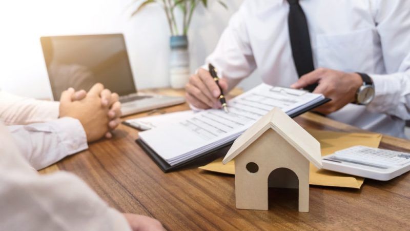 A Man Is Sitting at A Table with A Model House and A Clipboard — Linda Emery & Associates In Gosford, NSW