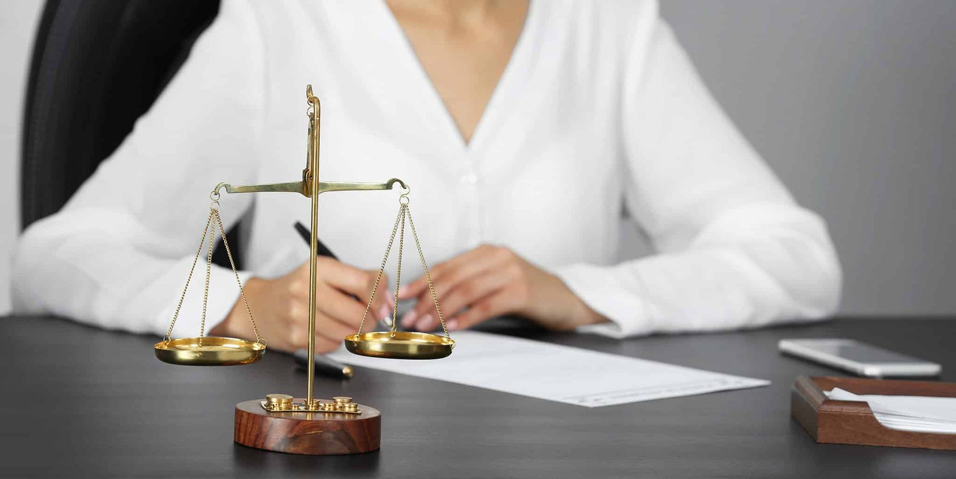 A Woman Is Sitting at A Desk with A Scale of Justice and Writing on A Piece of Paper — Linda Emery & Associates In Erina, NSW