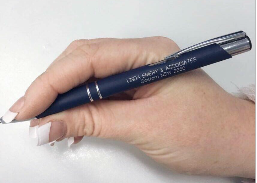 A woman's hand holding a blue pen with 'Linda Emery & Associates' written on it.