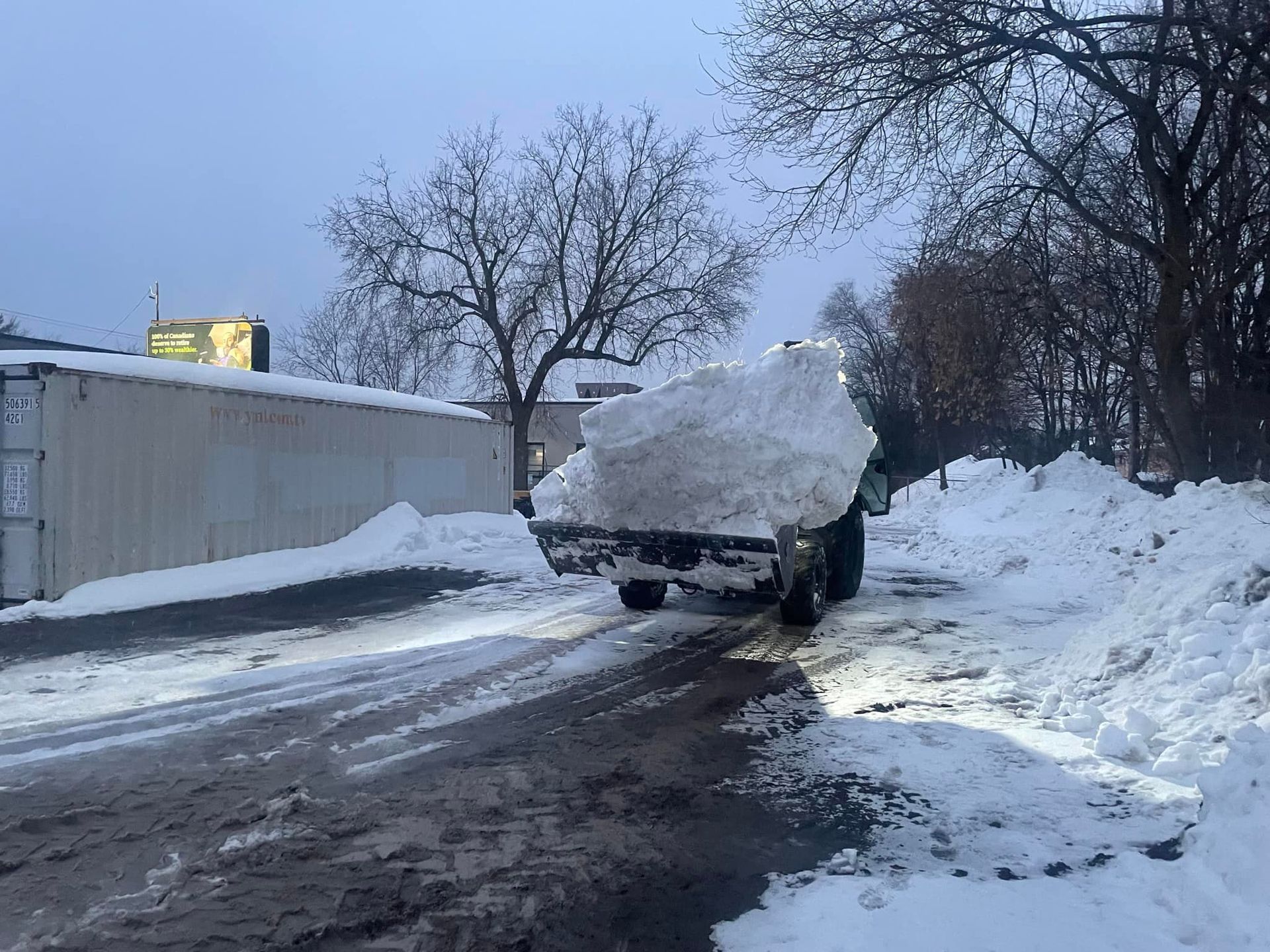 A truck heavily loaded with snow drives on a snowy road next to a building and trees.