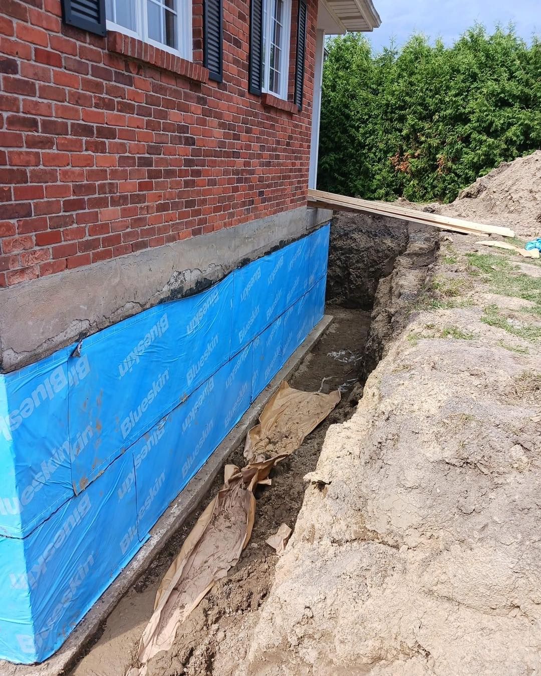 Foundation repair: Blue waterproof membrane installed on a brick house foundation, trench dug alongside.