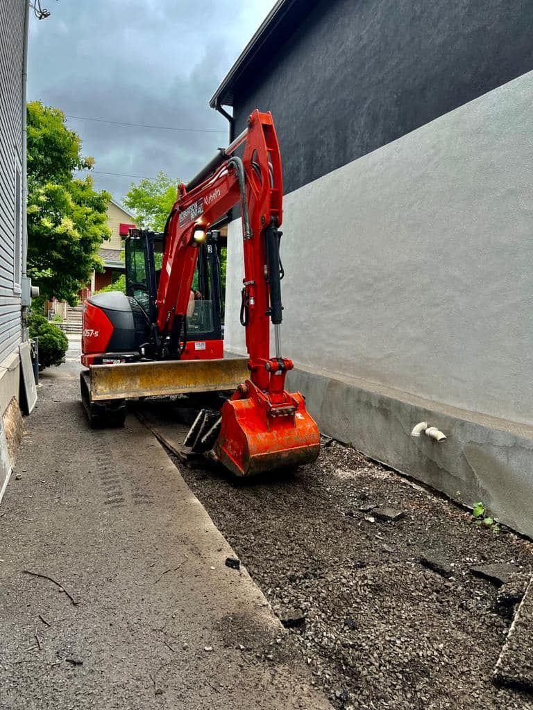 Red excavator digging in narrow alley between buildings.