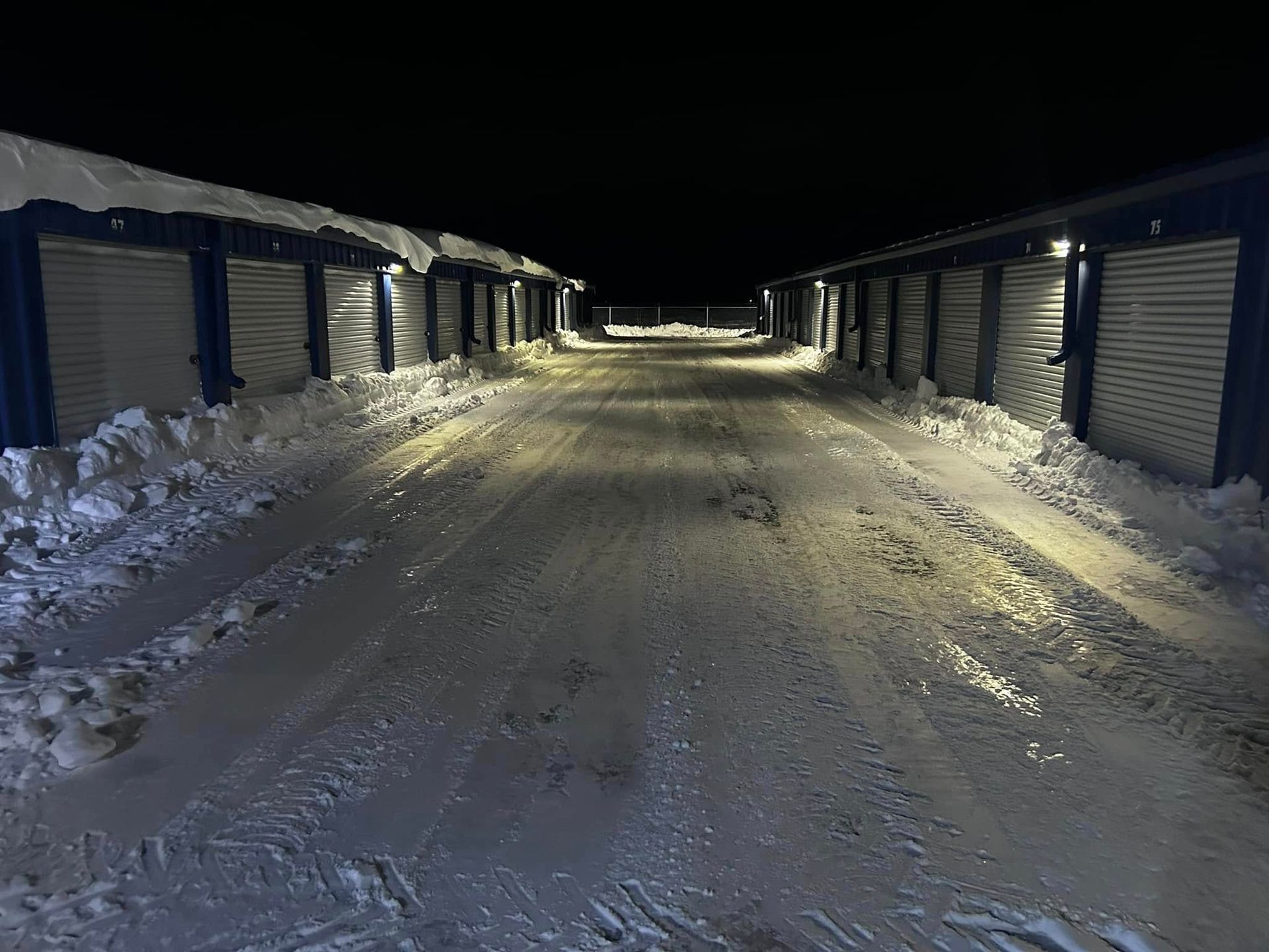 Storage units in a snowy lot at night, illuminated by lights.