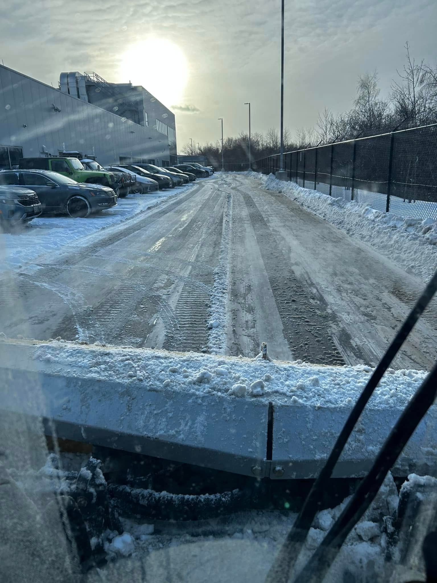 Snow plow clearing a parking lot on a cloudy day. Cars parked along the left, fence to the right.