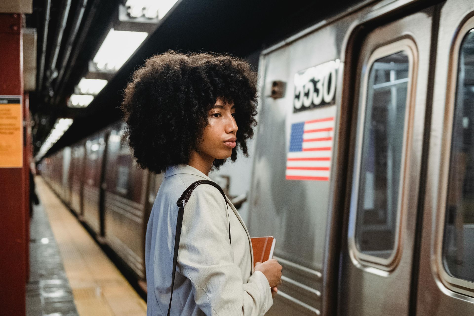 Woman waiting on a subway platform beside a silver train, holding a notebook.
