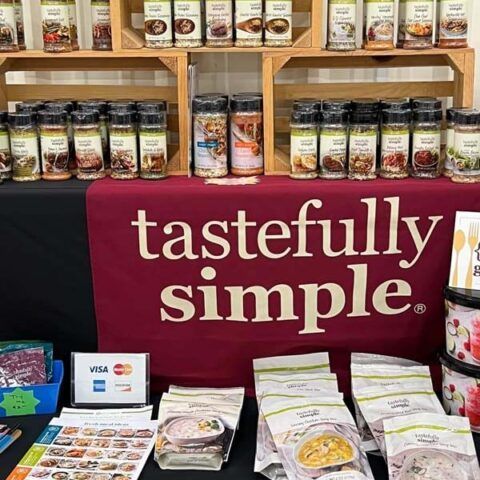 Booth display with maroon “tastefully simple” banner, shelves of packaged food, and product samples on a table