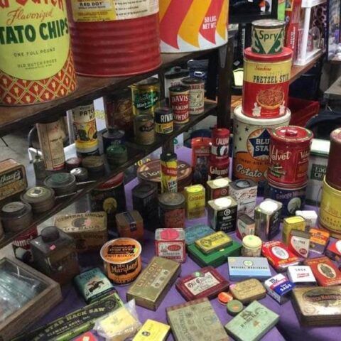 Assorted vintage cans and tins displayed on a crowded shelf and tabletop in a shop