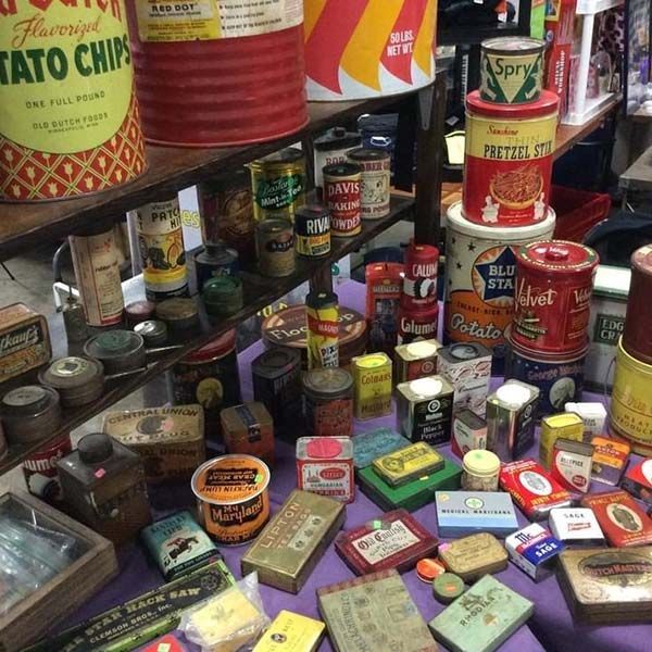 Assorted vintage tins and containers stacked on shelves and a table in a cluttered shop display.