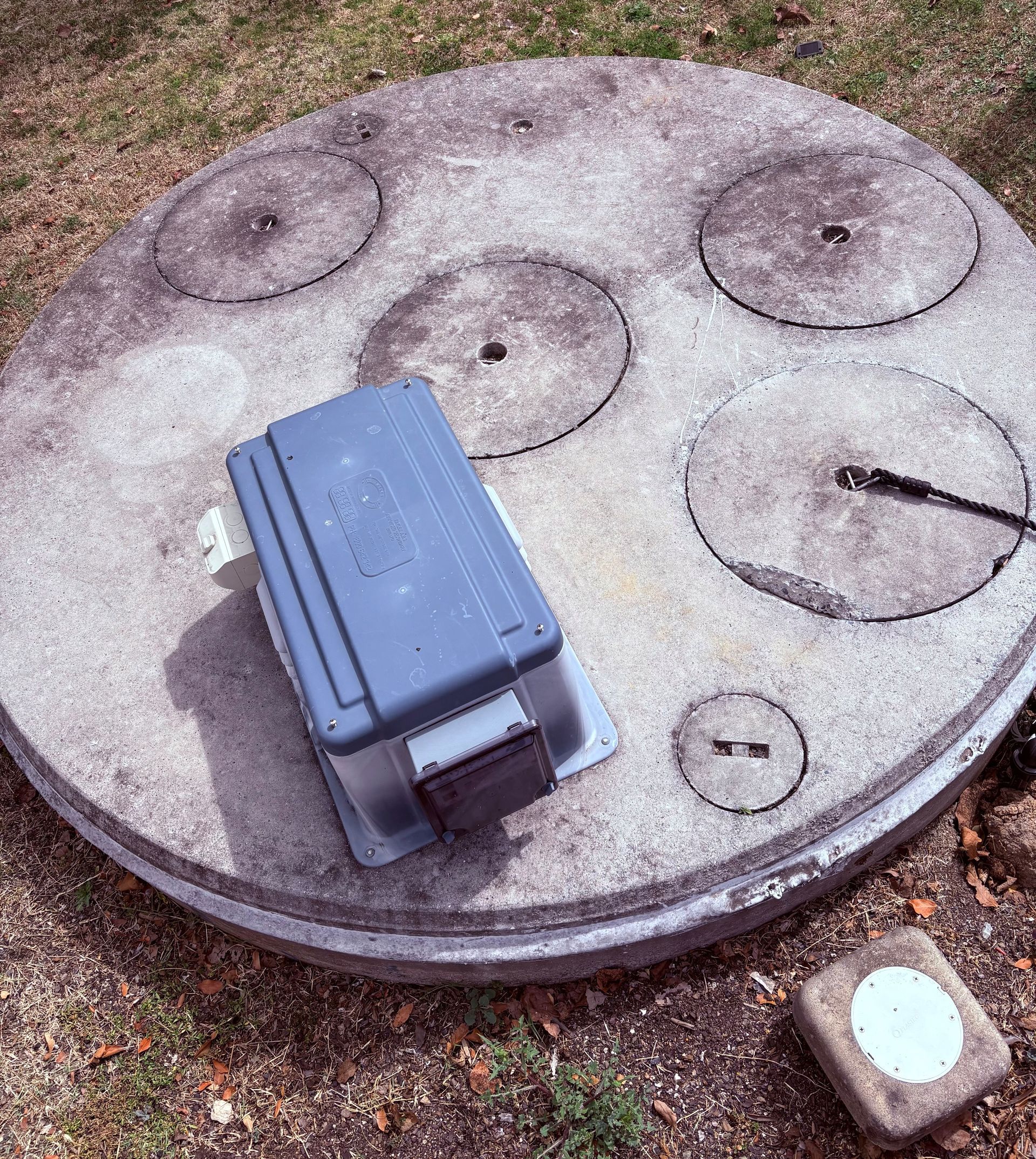 Construction worker near open manhole; orange safety vest, blue jeans, asphalt pavement, and equipment visible.