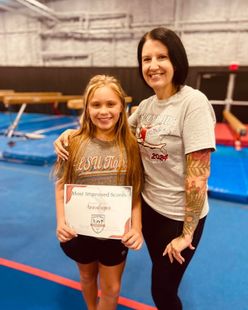 A girl with a certificate stands with a woman in a gym. Both smile, pose for photo.