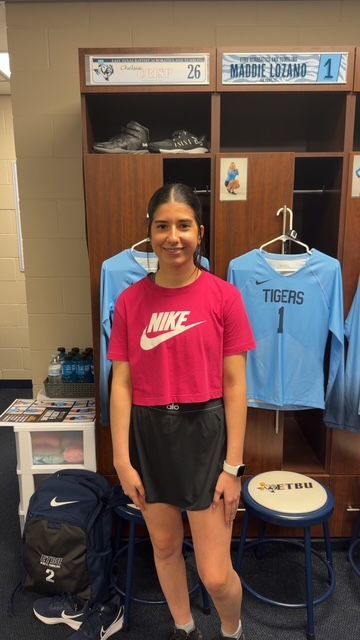 Woman in pink Nike shirt and black shorts stands in a locker room with team gear.
