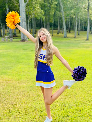 Cheerleader in blue and gold uniform holds pom-poms, smiling. Outdoors on a grassy lawn.