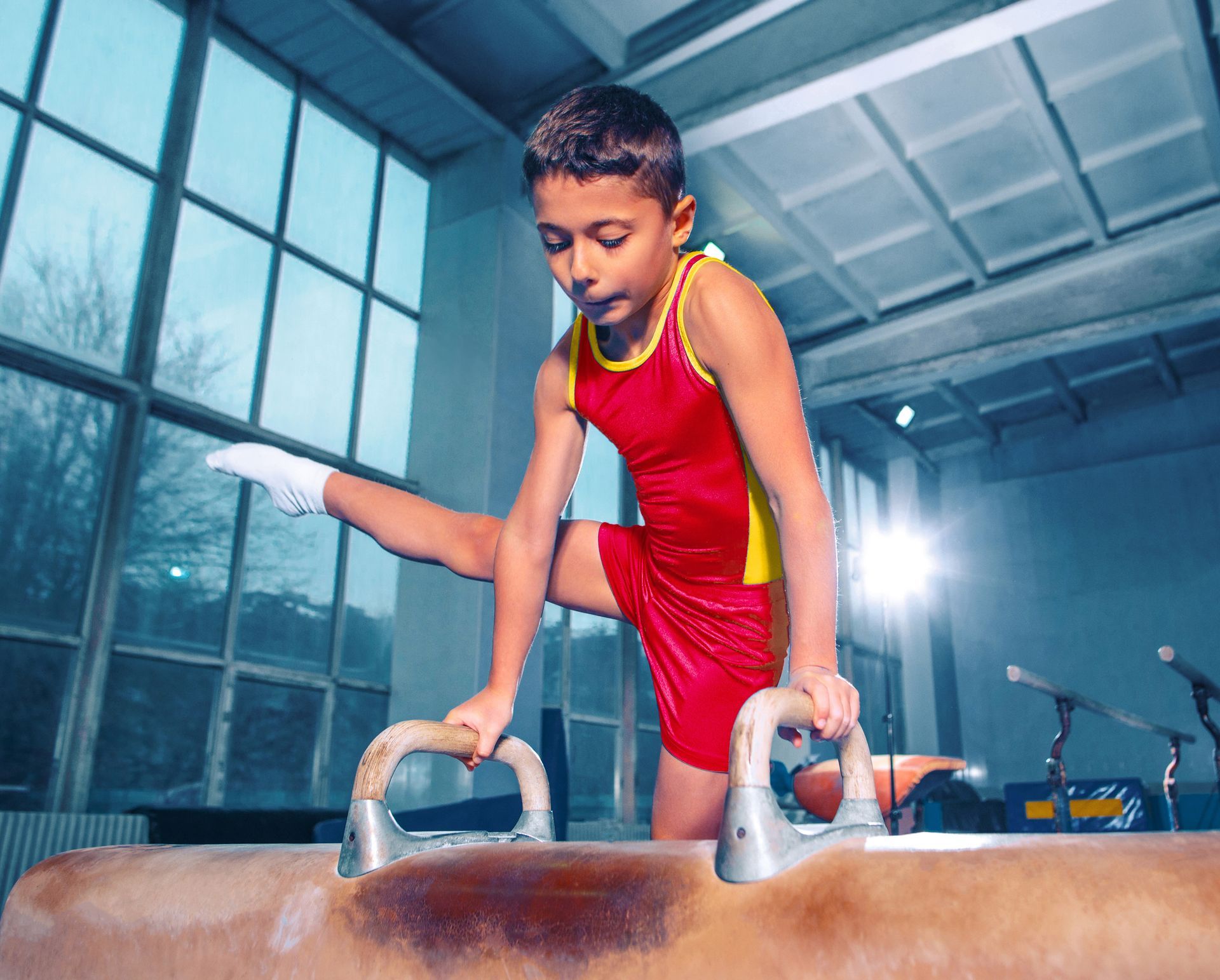 Young gymnast in red uniform on pommel horse, leg extended. Indoor gym setting.