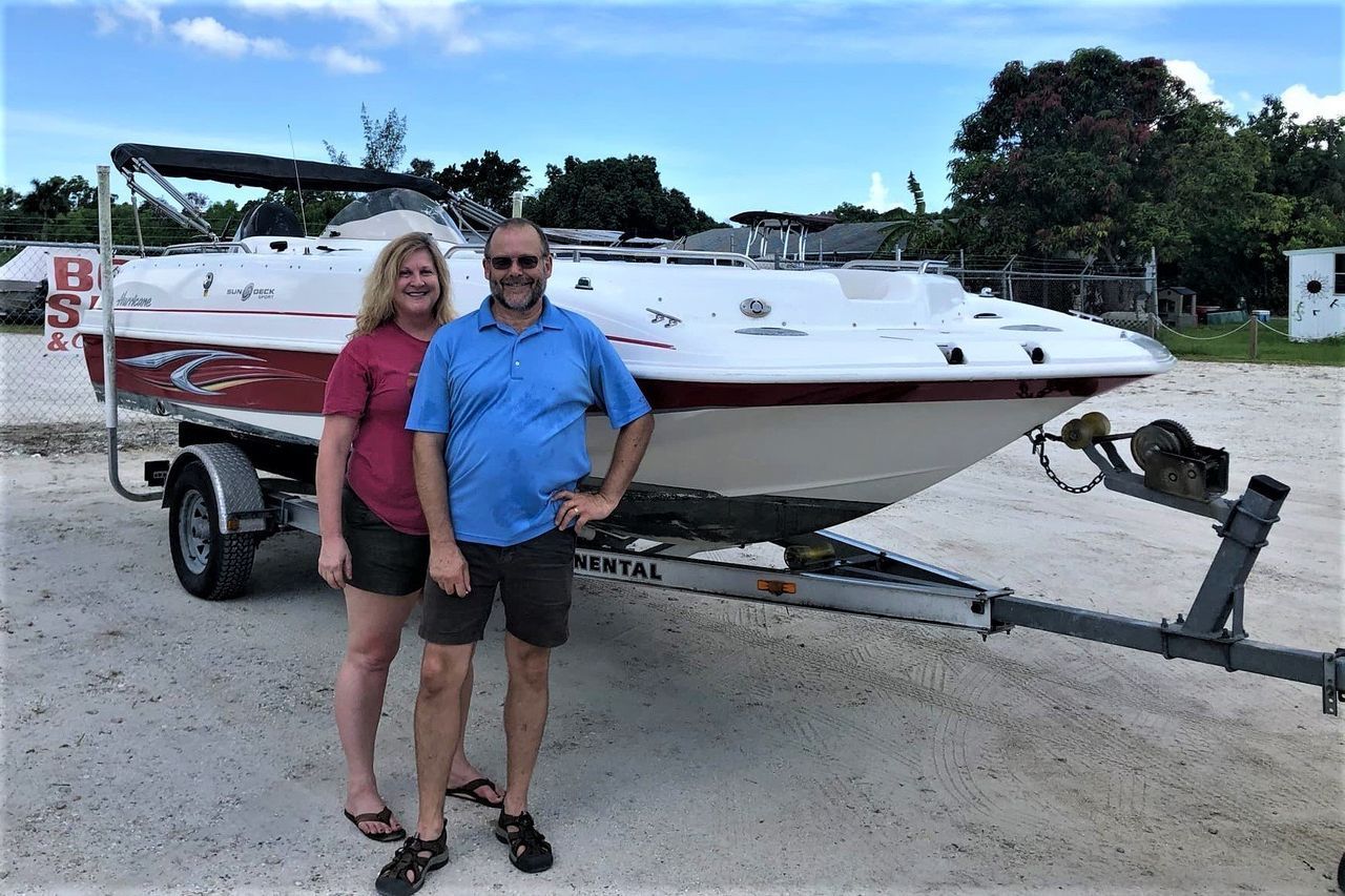 Two people standing beside a red-and-white boat on a trailer in a gravel lot