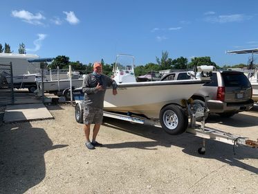 Man standing beside a silver boat on a trailer in a boatyard under a clear blue sky