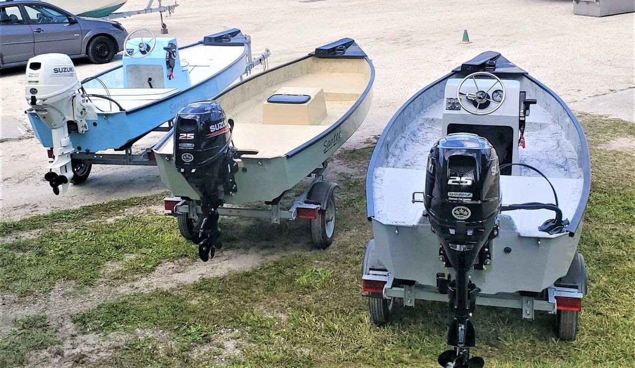 Three small boats on trailers, parked on a grass-covered lot, each equipped with an outboard motor.
