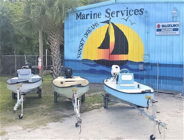Three small boats on trailers parked in front of a blue building with a mural of a sailboat.