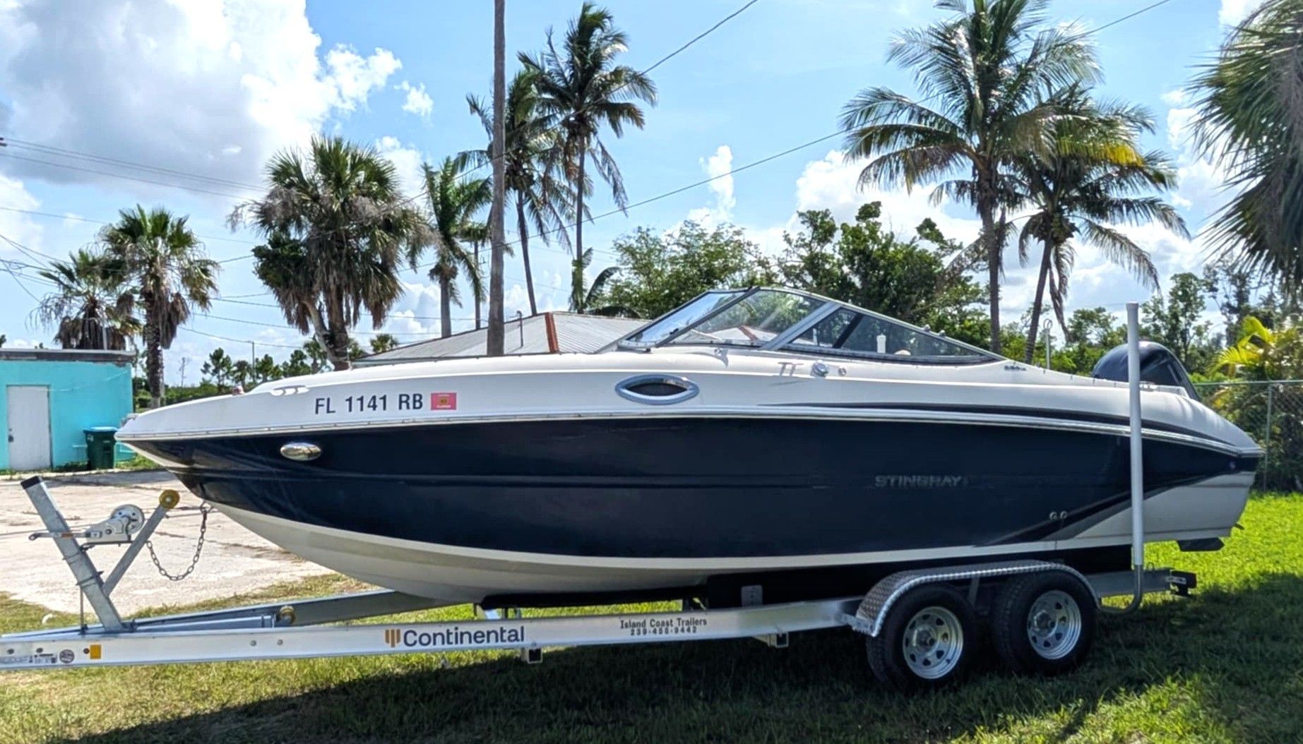 White and dark blue speedboat on a trailer in a sunny yard with palm trees in the background