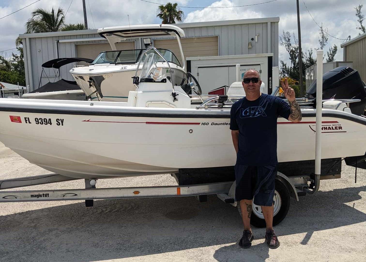 Man standing beside a white boat on a trailer in a gravel lot, with a building in the background