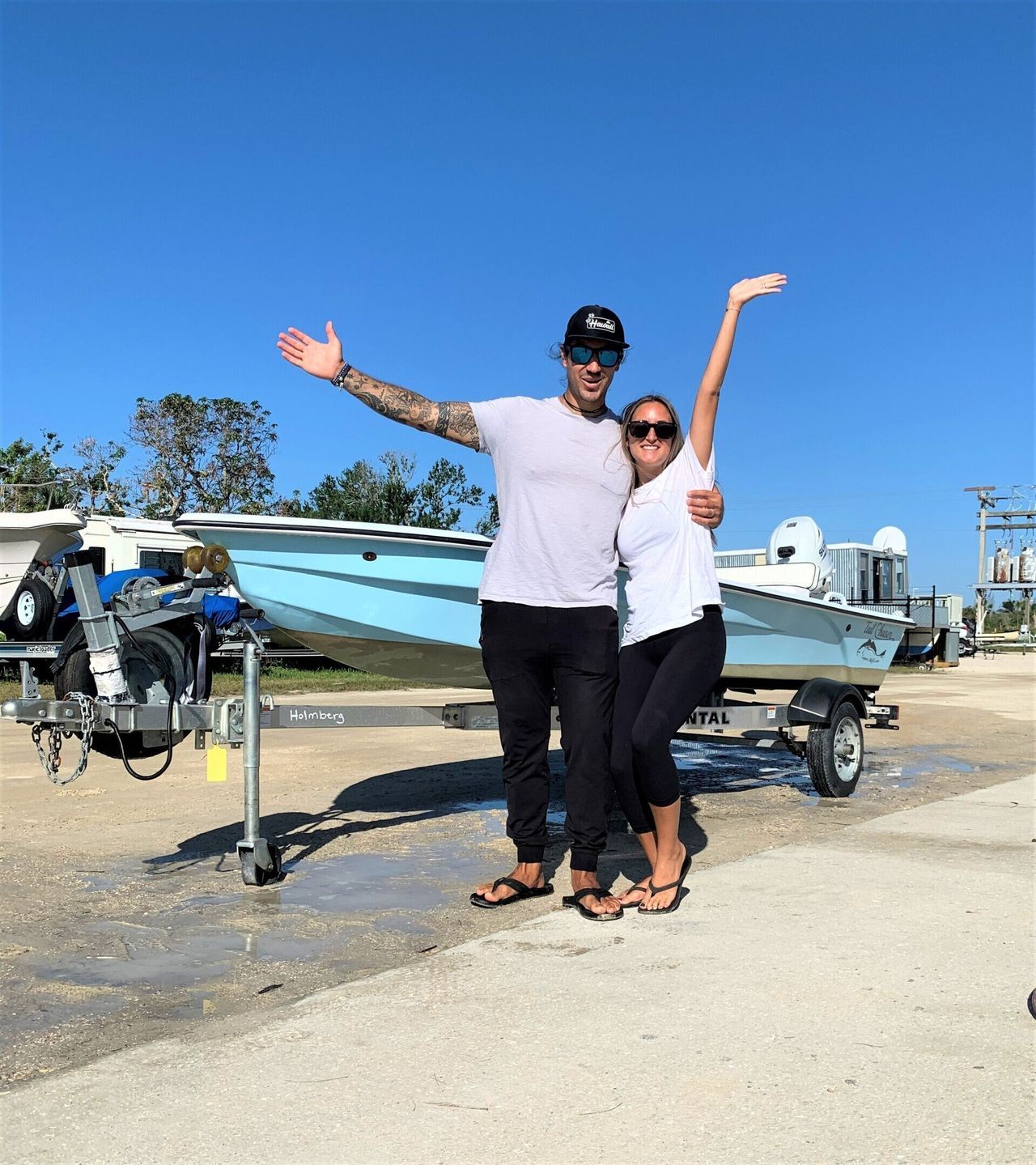 Two people smiling and posing beside a boat on a trailer under a bright blue sky