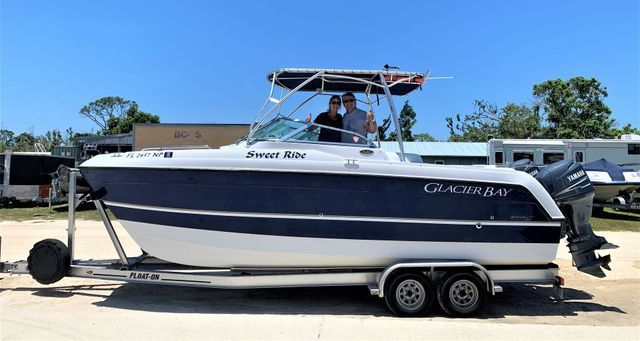 White and navy center-console boat on a trailer, with people aboard, parked outdoors under a clear blue sky.