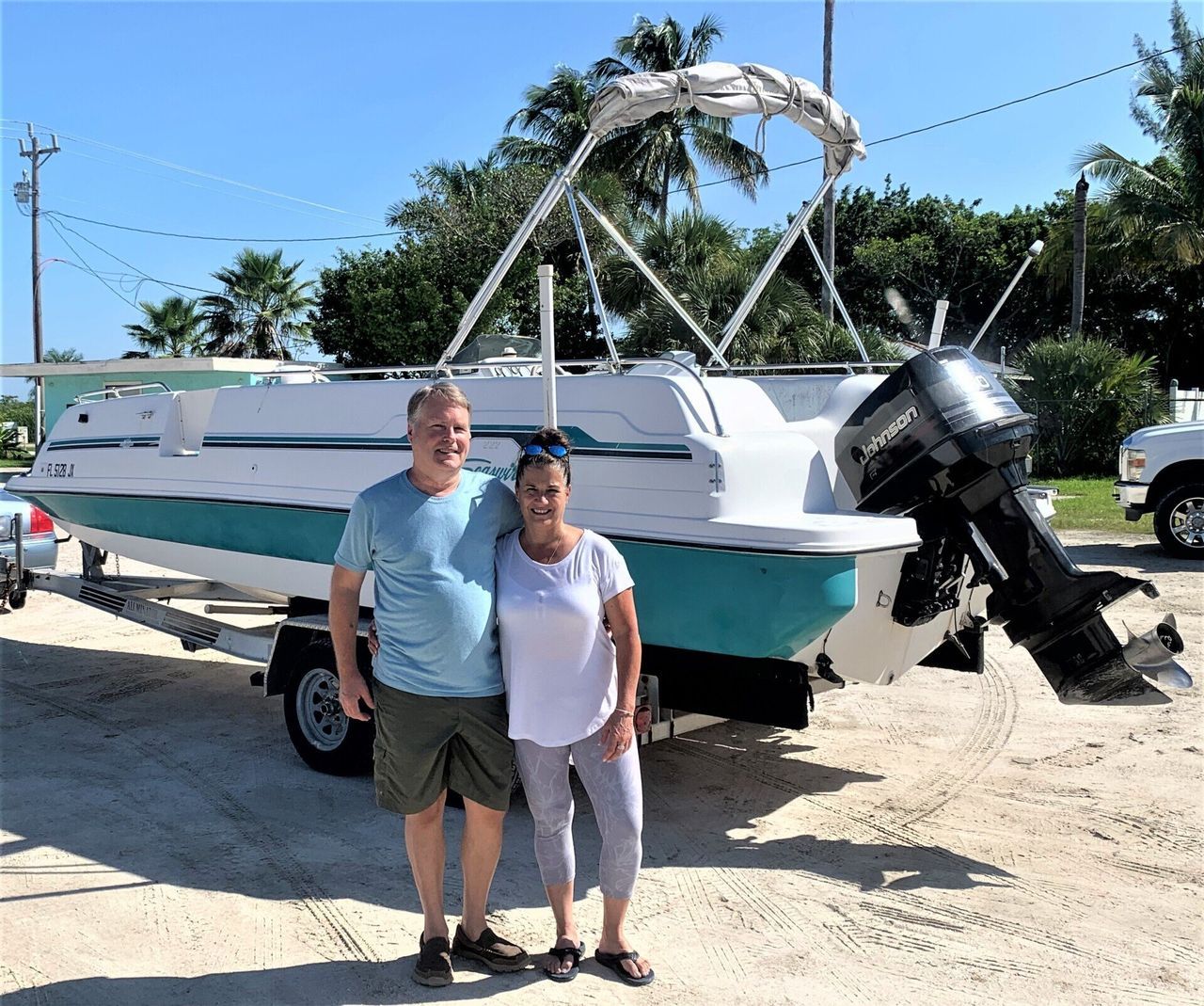 Couple posing in front of a teal-and-white boat with an outboard motor on a sandy lot