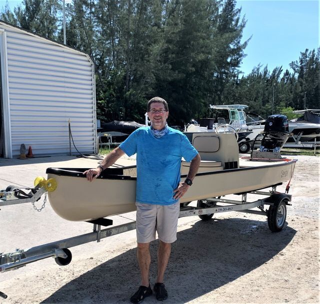 Man standing beside a tan boat on a trailer in a sunny outdoor yard with trees in the background
