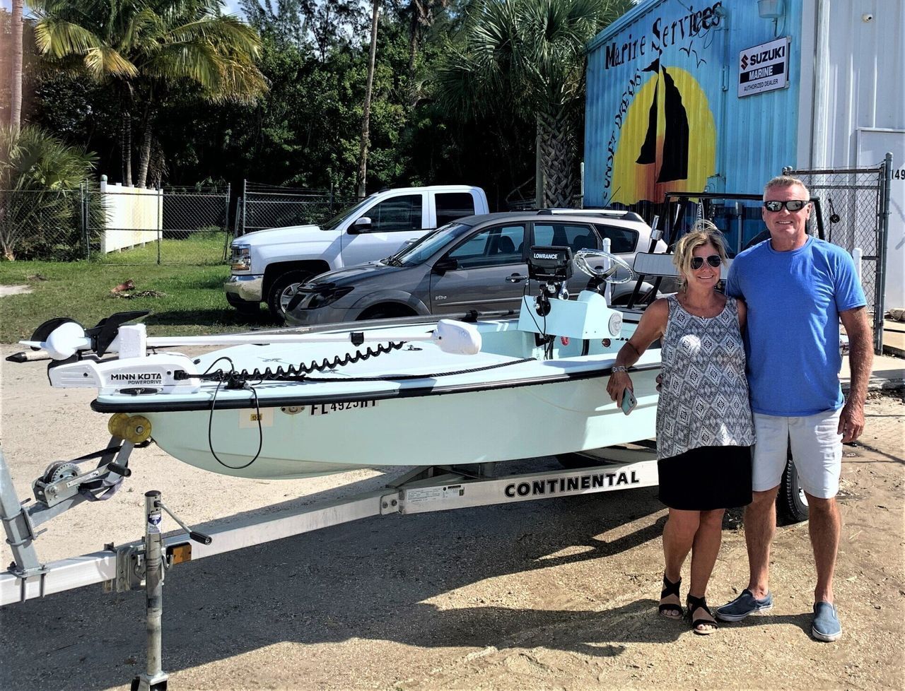 Two people posing beside a small white boat on a trailer at a marina, with vehicles and a building behind them.