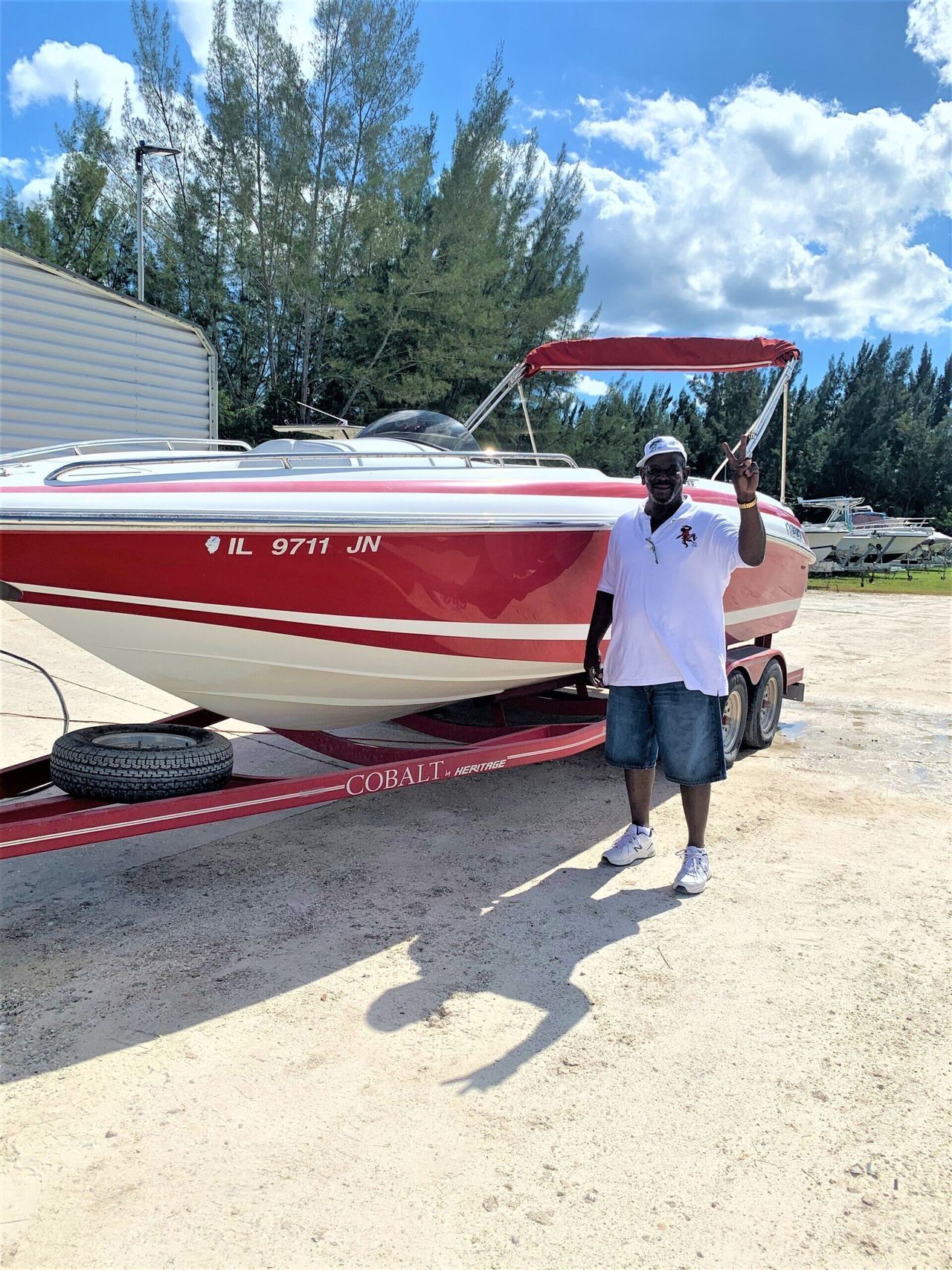 Man standing beside a red-and-white boat on a trailer in a sunny yard, holding up a paddle.