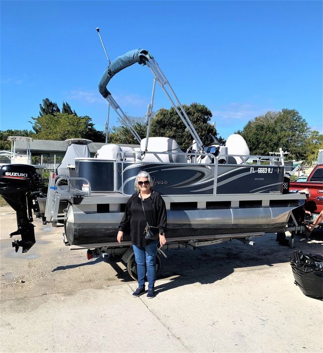 Person standing in front of a pontoon boat on a trailer in a sunny parking lot