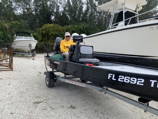 Two people on a black boat trailer at a gravel marina, one wearing a yellow shirt and white hat