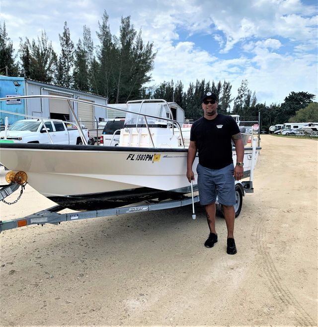 Man standing beside a white boat on a trailer in a sandy lot under a partly cloudy sky